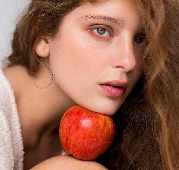 Young woman with long brown hair and her face is shine and glorious. A red apple rests under her chin, conveying a serene and natural mood. Soft lighting enhances her features.